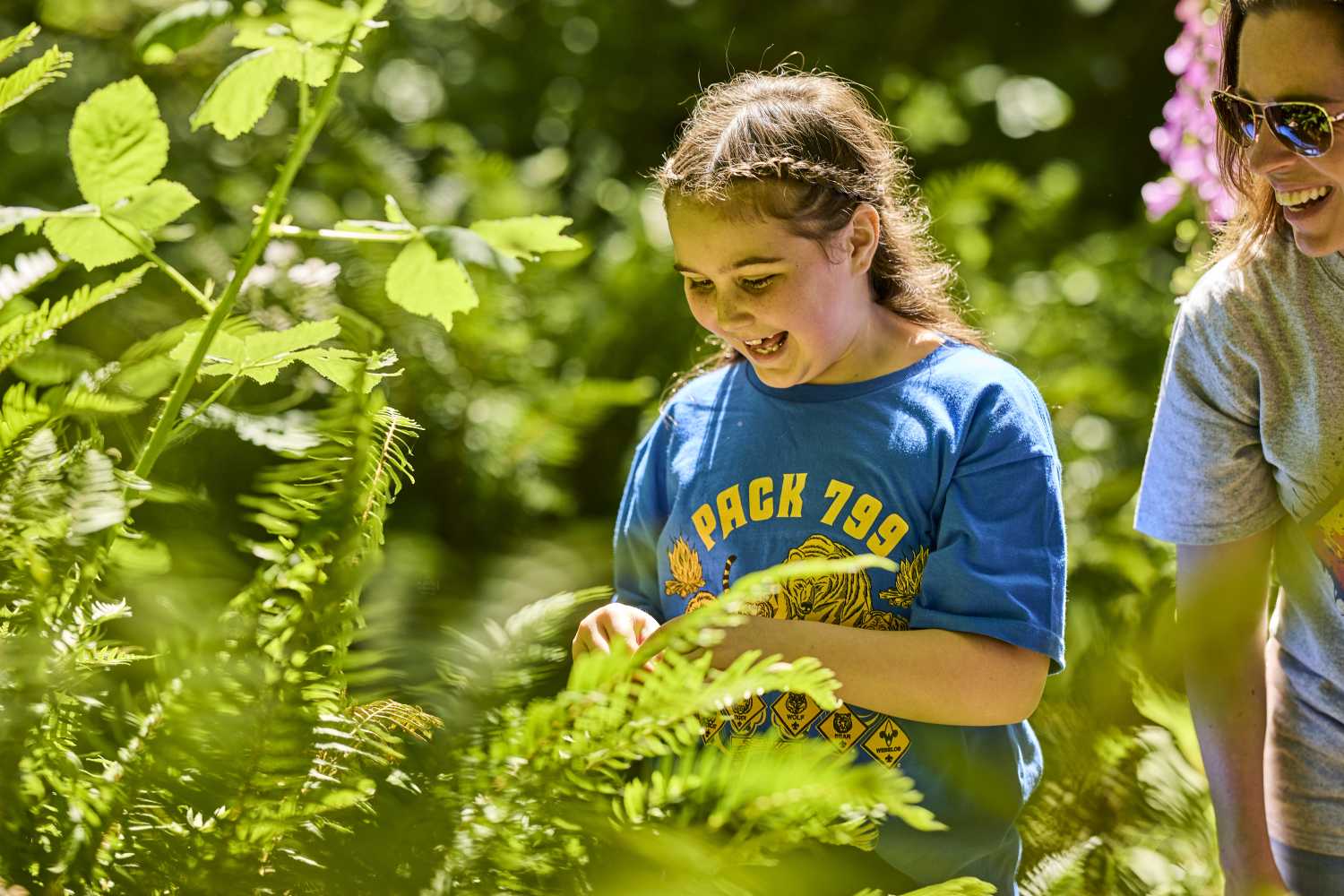 A young girl smiles brightly while looking down at a plant in the woods
